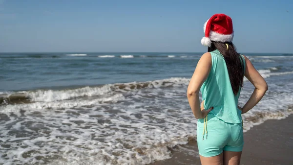 年轻女子戴着圣诞老人的帽子 站在海滨 在阳光灿烂的日子里遥望远方的背影 头戴圣诞礼帽的黑发成年人在海滨度假庆祝新年 — 图库照片
