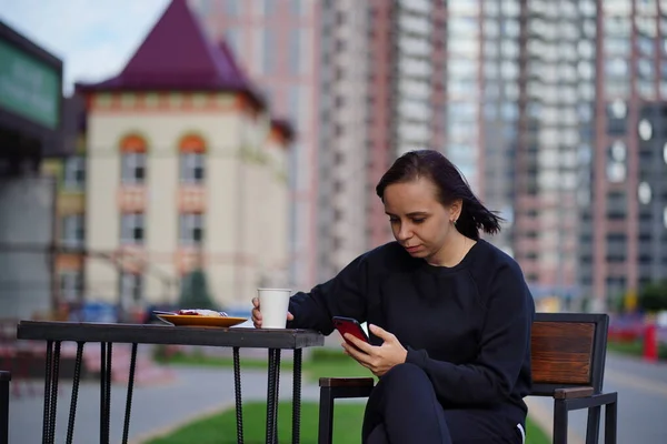 A young woman uses her cell phone outdoors. Beautiful woman sitting on a chair, holding a white mug with a hot drink. Concept of leisure and leisure.