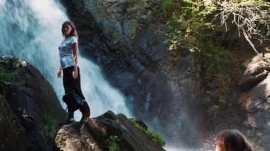 Kuzey Osetya, Rusya 1 Eylül 2020: Yazın güçlü bir şelalenin yanında poz veren genç bir kadın. Dişi gezgin, taşın üstünde büyük bir su yolu kenarında dikilirken fotoğraflandı..