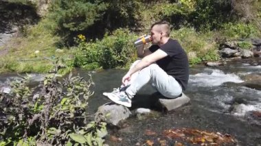 Genç adam dağ nehrindeki taşlara oturur ve termo bardaktan kahve ya da çay içer. Erkek turist, dağlarda aktif bir yürüyüş yaptıktan sonra dinleniyor ve güzel bir çevre manzarasının tadını çıkarıyor.