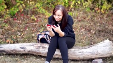 Kütüğün üzerinde oturan, cep telefonuna göz atan ve kahve içen genç bir kadın. Yetişkin esmer, ormanlık alanda akıllı telefon ve sıcak içeceklerle dinleniyor.