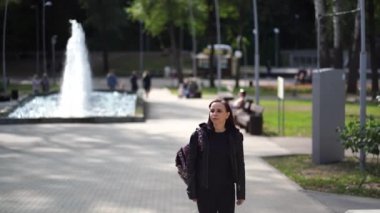 Sırt çantalı, deri ceketli güzel bir bayan şehir parkında yürüyor. Yetişkin esmer, güneşli bir havada sokakta geziniyor.