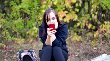 Kütüğün üzerinde oturan, cep telefonuna göz atan ve kahve içen genç bir kadın. Yetişkin esmer, ormanlık alanda akıllı telefon ve sıcak içeceklerle dinleniyor.