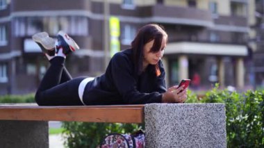 Genç bir kadın, dışarıda bir bankta uzanmış, bacakları yukarda ve cep telefonunu karıştırıyor. Akıllı telefon kullanan bir kadın bir apartmanın avlusunda internet kullanıyor..