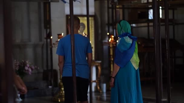 Mężczyzna i kobieta w chustce modlą się w świątyni. Parafianie stoją przed ikoną i modlą się z całego serca w kościele.. — Wideo stockowe