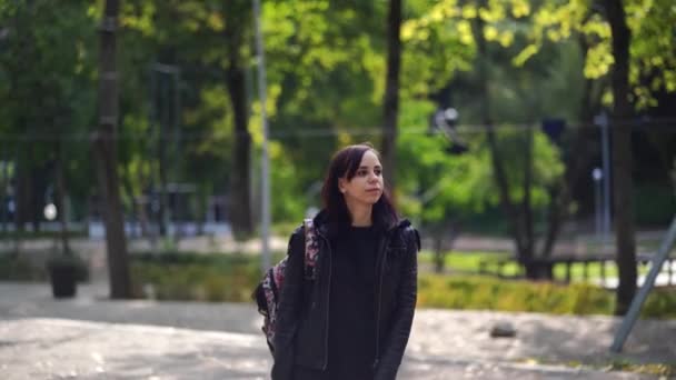 穿着皮夹克背着背包在城市公园散步的年轻漂亮的女人 在阳光明媚的天气里 黑发成年人在街上漫步 — 图库视频影像