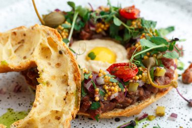 Çiğ Bıldırcın yumurta sarısı ile sığır tardara dolu sağlıklı taze pişmiş simit, turşu kapanlar, mikroyeşillik, kurutulmuş domates, hardal ve kesilmiş soğan. Beyaz plaka üzerinde hizmet vermiştir. Yakın görünüm