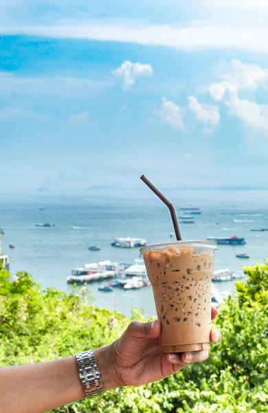 Hand holding iced coffee and sea with many boats at Pattaya in Thailand.