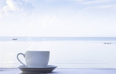 Tahta bir masanın üstünde çay tabağı olan beyaz bir fincanda sıcak kahve. Arka plan bulanık tekne ve deniz..