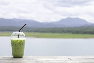 Masanın üzerinde bir bardak soğuk yeşil çay. Arka plan bulanık su ve dağ manzarası..