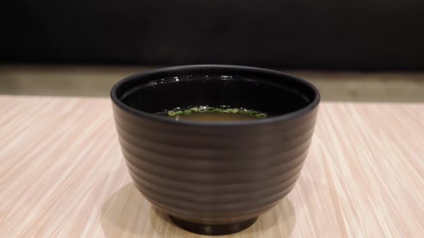 White smoke floating from the broth with spring onions in a black cup on wooden  table.