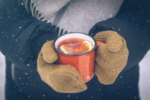 Mulled wine in a red cup in women's hands mittens. Hot winter drink outdoor in snowy winter forest. Christmas background with snowflakes
