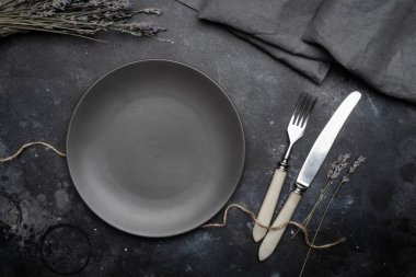 Boş gri plaka (seramik) üzerine koyu gri bir arka plan ile bıçak ve çatal, lavanta ve peçete bir buket ile dekore edilmiştir. Gri minimalist kavramı. Kopya alanı.