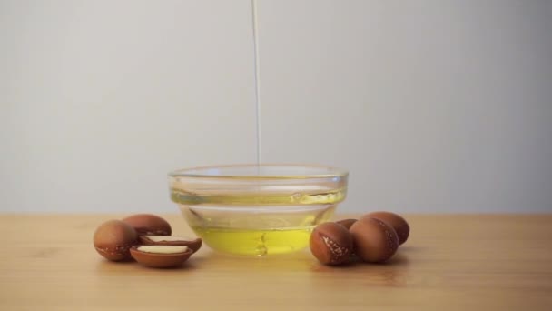 Aceite de argán vertiendo en un recipiente de vidrio con semillas de argán sobre un fondo blanco. Concepto de cosméticos a base de aceite de argán. — Vídeo de stock