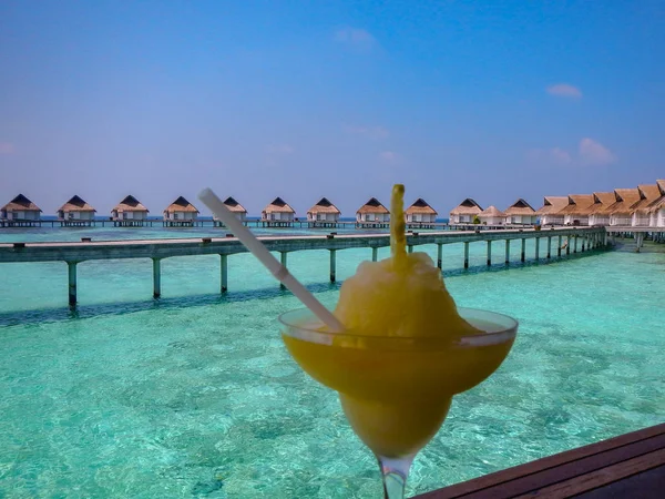 Huzurlu bir yaz tatili için yürüyüş yolu ve sazdan kulübeleri ile köpüklü bir lagün bakan ahşap güverte tropikal bir ada Resort dondurulmuş tatlı veya kokteyl