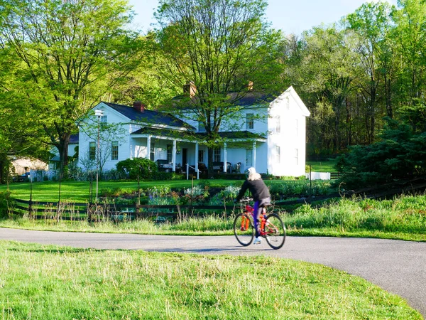 Kadın yolun beyaz bir çiftlik yapraklı yeşil ağaçlar arasında geçmiş bir viraj etrafında Bisiklete binme