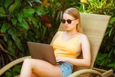 Bir şezlong üzerinde yalan ve bir dizüstü bilgisayar üzerinde çalışan güneş gözlüklü kız