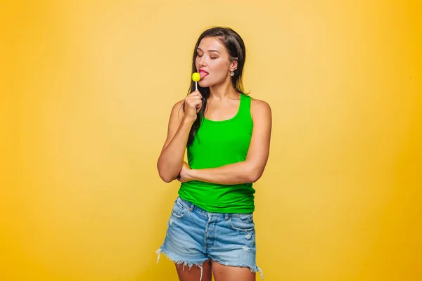 Девушка на жёлтом фоне в зелёной футболке ест леденец — стоковое фото