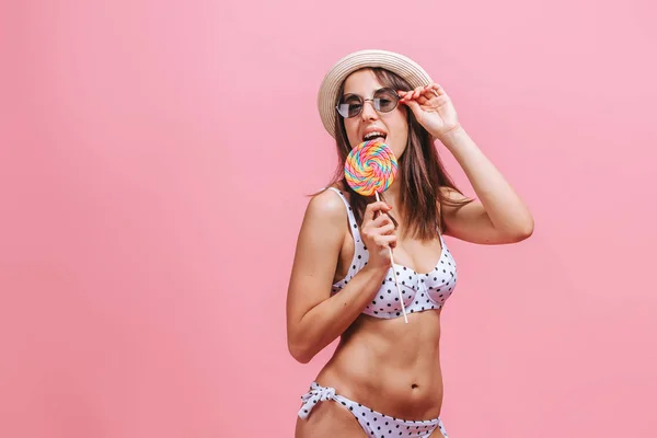 Chica comiendo piruleta en gafas de sol traje de baño y sombrero en la espalda rosa — Foto de Stock