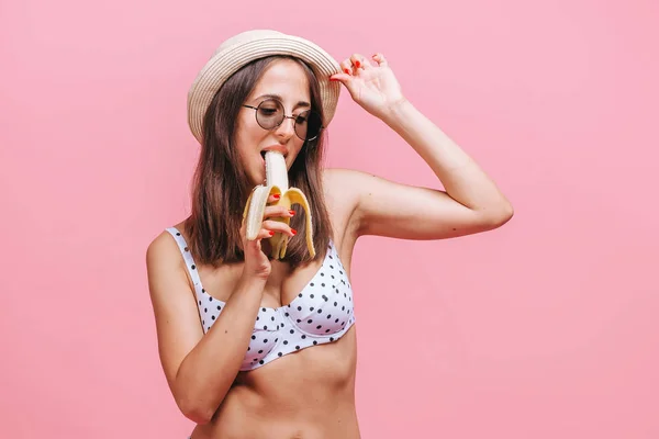 La chica se come un plátano en traje de baño, sombrero y gafas de sol en un pi — Foto de Stock