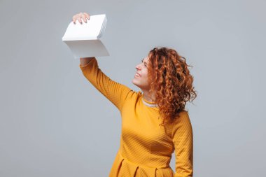 Açık gri bir arka plan üzerinde beyaz bir kitap tutan kızıl saçlı kız.