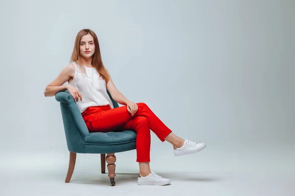 Chica en pantalones rojos sentada en una silla sobre un fondo gris claro — Foto de Stock
