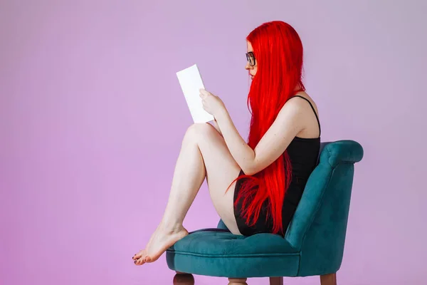 Kırmızı uzun saçlı kız bir sandalyede oturan ve bir kitap okuma