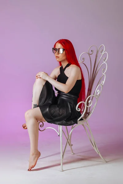 Chica con el pelo largo rojo con gafas y una falda de cuero se sienta —  Fotos de Stock