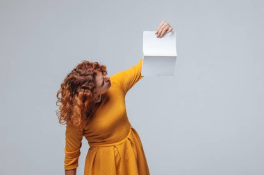 Açık gri bir arka plan üzerinde beyaz bir kitap tutan kızıl saçlı kız.