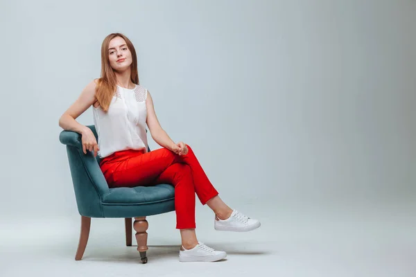 Chica en pantalones rojos sentada en una silla sobre un fondo gris claro — Foto de Stock