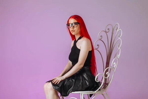 Ragazza con i capelli lunghi rossi indossando occhiali e una gonna in pelle siede — Foto Stock