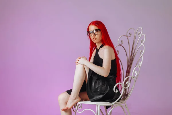 Girl with red long hair wearing glasses and a leather skirt sits — Stock Photo, Image