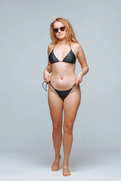 Chica en un traje de baño negro y gafas de sol en un fondo gris claro — Foto de Stock