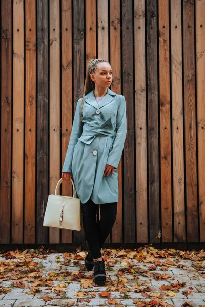 가을 코트를 입고 가방을 들고 나무 뒤 뜰에 서 있는 소녀 — 스톡 사진