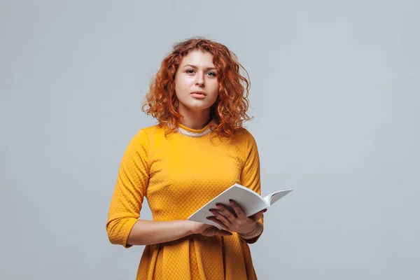 Chica pelirroja leyendo un libro blanco sobre un fondo gris claro . —  Fotos de Stock