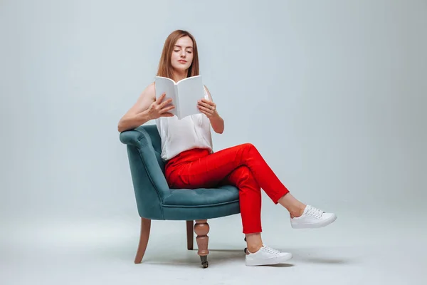 Chica en pantalones rojos sentada en una silla y leyendo un libro con una w —  Fotos de Stock