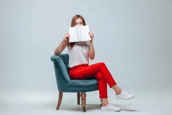 Chica en pantalones rojos sentada en una silla y leyendo un libro con una w —  Fotos de Stock