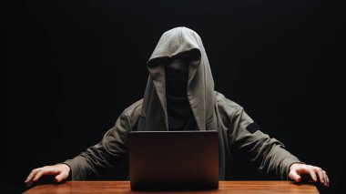 Kapüşonlu suratsız bir bilgisayar korsanı siyah bir sırt çantasına dizüstü bilgisayarı takmış.