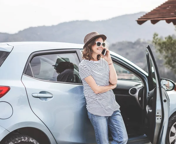 Young beautiful female travel girl in hat and sunglasses speaks by mobile phone on the background of her car while traveling in the mountains, renting and car service