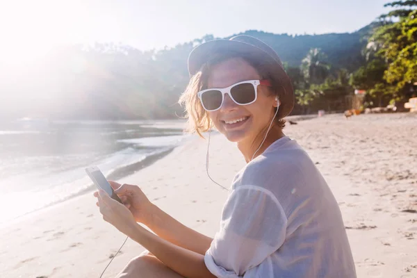 若くてきれいな女性流行に敏感な旅行者手 太陽の下でビーチで携帯電話とヘッドフォンで — ストック写真