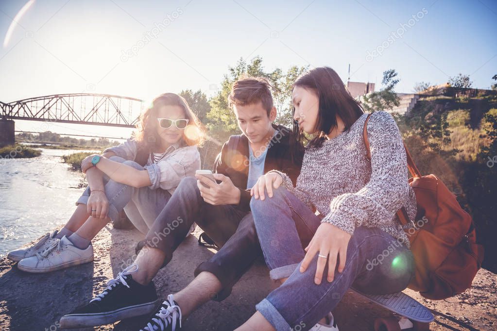 Company school friends teens of different ethnic nationalities sit outside, relax, have fun with smartphone, in the rays of the setting su