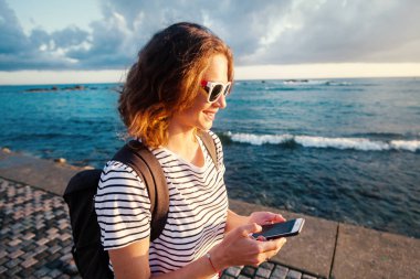 Bir bağlantı noktası sahil yürüyüş ve izlerken kitle iletişim araçları memnun üstünde bir akıllı telefon ile deniz günbatımı arka planda gülüyorsun mutlu rahat kadın
