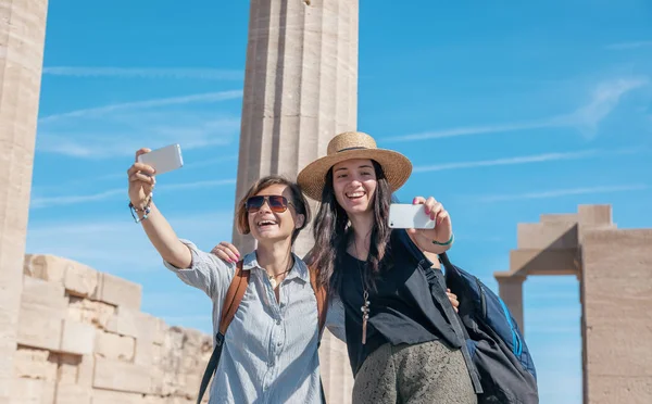 Two young beautiful happy woman female travel girlfriends doing selfie on the background of the sea and ancient ruins in Greece