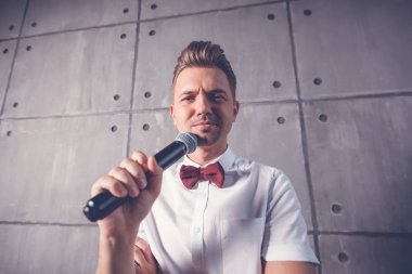 Genç çekici komik neşeli duygusal biri beyaz bir gömlek ve bir kelebek bir mikrofon ile gri bir arka plan üzerinde onun elinde winks