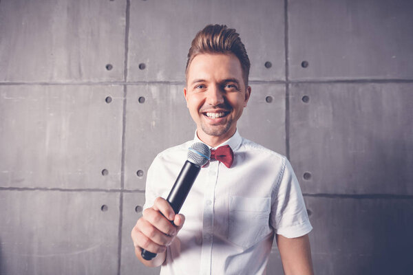 A young attractive funny joyful emotional man winks in a white shirt and a butterfly with a microphone in his hands, on a gray background