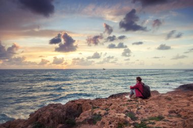 Genç kadın gezgin kış deniz kıyısında oturur ve parlak renkli gün batımını izler