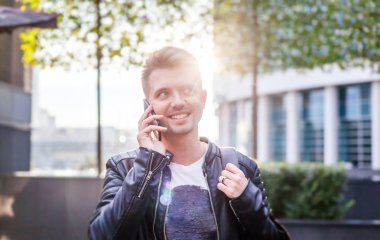 Çekici şık genç adamla bir cep telefonu ile bir deri ceketli bir smartphone sabah güneşi şehrin sokaklarında onun elinde
