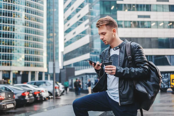 革のジャケットの大規模な近代的な都市の背景に彼の手でスマート フォンとテイクアウト コーヒーをスタイリッシュな若い男は魅力的 — ストック写真