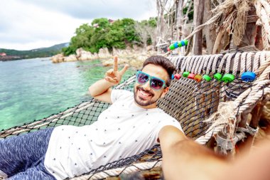 Çekici yakışıklı genç Latin bey, karanlık bir hamak tatil sakallı selfie Smartphone cep telefonu ile deniz keyfi hayat karşı kılıyor