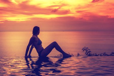 Bikinili genç ince kadın sonsuzluk havuzu kenarında parlak bir gün batımı arka plan üzerinde tatil beldesi oturur
